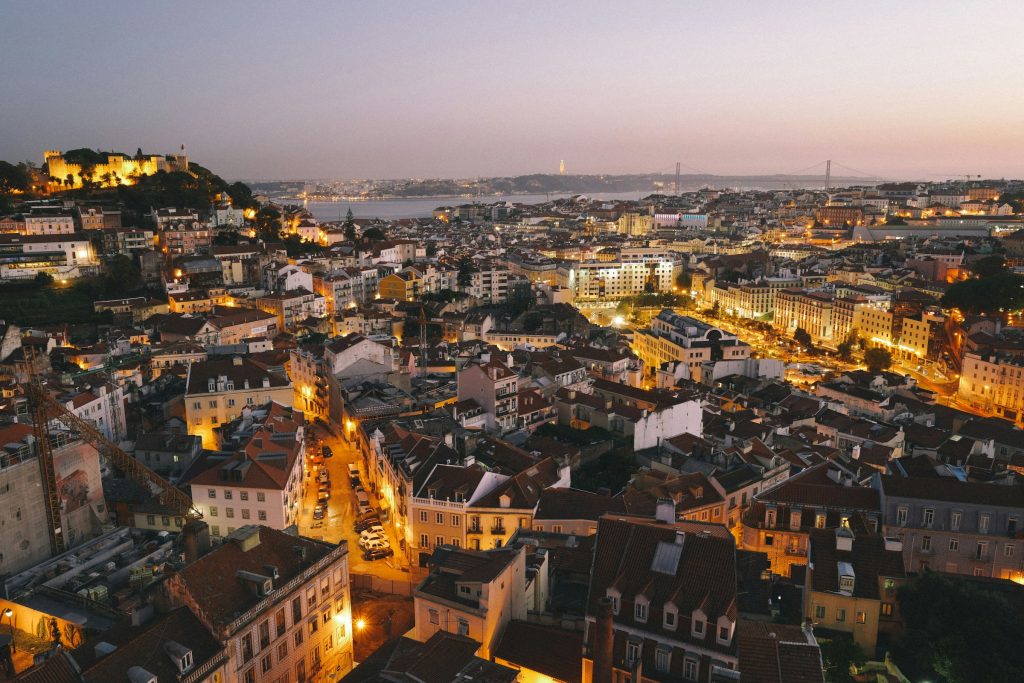 Twilight panorama over Lisbon’s hillside rooftops and the Tagus River, with São Jorge Castle lit up on the left and the 25 de Abril Bridge in the distance
